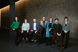 The seven Theatertreffen jury members stand or sit in front of a pebble wall and look into the camera.