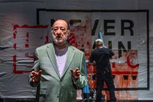 An elderly man explains something with a serious expression while someone in the background works on a blood‑smeared poster