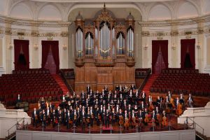 Die Musiker*innen eines Orchesters stehen in einem Konzertsaal vor einer großen Orgel.