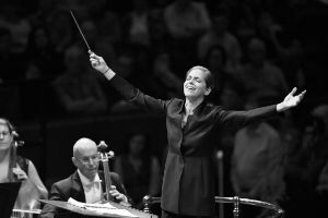 A conductor spreads her arms in front of the orchestra, the musicians bow their violoncelli and the audience can be seen in the background.