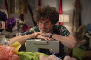 A man is sitting in his sewing studio with both arms resting on his sewing machine.