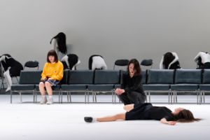 Eine Person in gelbem Pullover sitzt auf einer Stuhlreihe und schaut auf eine andere Person, die auf dem Boden liegt.