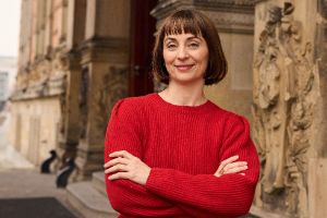 Jenny Schlenzka, dressed in a red costume, stands smiling with her arms folded in front of the entrance to the Gropius Bau.