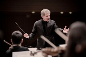 A conductor stands in front of musicians with outstretched arms and a conductor's baton in the right margin.