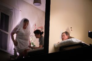 Eine Frau horcht an einer hellgelben Wand, hinter der eine Braut angekleidet wird.