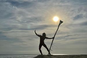 A silhouette of a person against the light standing on the beach, stretching one arm towards the sky.