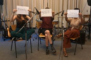 Three people sit on chairs in a music studio and hold pieces of paper with the words Ready SET and Jazz in front of their faces.