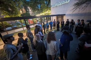 Stipendiat*innen des Internationalen Forum stehen abends im Kreis vor der Seitenbühne der Berliner Festspiele. Viele haben ein Glas Wein in der Hand und hören einer sprechenden Person zu.