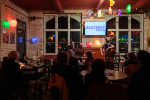 Publikum sitzt an Tischen auf Stühlen mit Blick zum Konzert der Musiker*innen Elin Forkelid am Saxofon, Anton Jonsson am Schlagzeug und Vilhelm Bromander am Kontrabass.