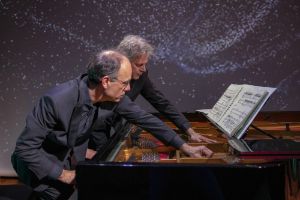 Portrait of Andreas Grau and Götz Schumacher, standing behind an open grand piano, whose strings they are fingering.