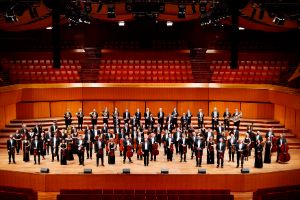 Musicians from a large orchestra stand with their instruments in a concert hall and look into the camera.