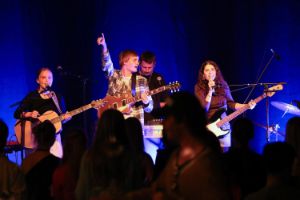 Four young musicians perform on a blue-lit stage with guitars and microphones.
