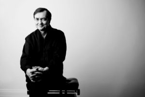 Man sits on a piano stool facing the viewer in front of a white background