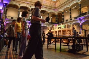 Vier Menschen sehen am linken Bildrand aufgereiht mit Tischtennisschlägern in der Hand im Lichthof des Gropius Bau und schauen anderen beim spielen zu.