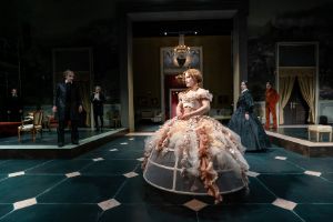 A young woman in a hoop skirt stands in a salon, with more people in historical costumes in the background