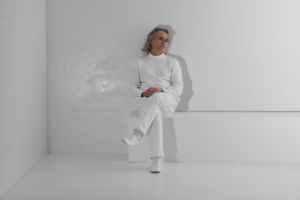 A man dressed in white sits with his legs crossed on a white bench inside a white cube