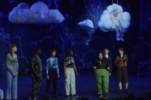 A group of young people stand on a blue-lit stage. Clouds hang from the ceiling.