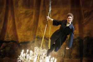 A woman in a blazer‑coat stands smiling on a swinging chandelier