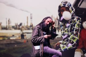 Arne Vogelgesang sits on a stage and looks into a tablet. In the background is a screen showing a young man with a face mask.