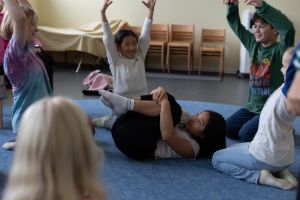 Children sit in a circle and stretch their arms in the air. In the centre of the circle, another child is curled up into a little package.