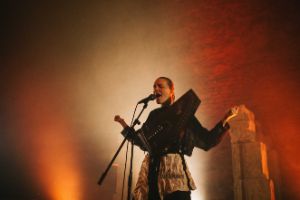 A photo of a person singing into a microphone and carrying an autoharp in front of their stomach.