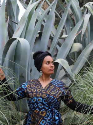 A Woman of Colour stands in front of large plants and looks into the distance