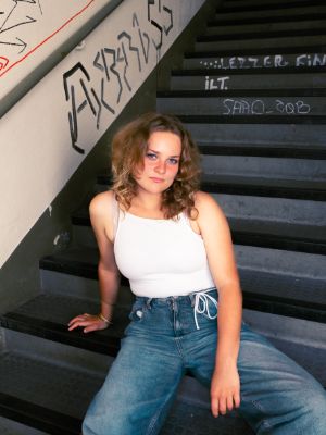 Eine weibliche Person sitzt auf einer  Treppe und schaut selbstbewusst in die Kamera. Sie trägt Jeans und ein blaues Top.