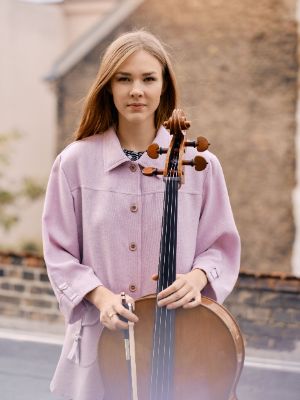 Valerie Fritz mit einem Cello in der Hand vor einem Backsteinhaus