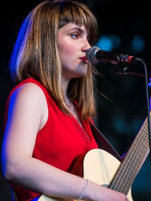 Kamai Melon mit ihrer Gitarre auf der Bühne. Sie trägt ein rotes Kleid und singt in ein Mikrofon.