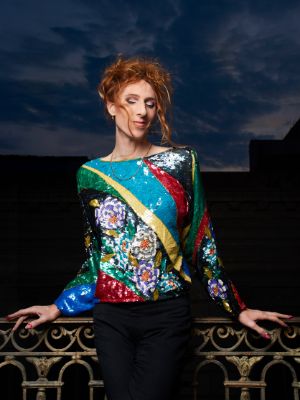 Nina Laisné wears a colourful top and leans against a curly railing with her eyes closed.