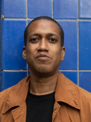 A man in front of a tiled blue wall looks into the camera.