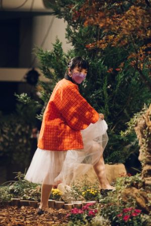 A female read person in a white tulle skirt and a red kimono, lifting one leg and holding her skirt with her hands. She is in front of a garden landscape, which is located in a hall.