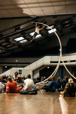 A female read acrobat is hanging from a concave scaffold. An audience is sitting below her.