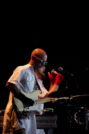 Zwei männlich gelesene, in Weiß gekleidete Personen während eines musikalischen Auftritts. Die Person im Vordergrund spielt Gitarre, die Person im Hintergrund hält ein Mikrofon.