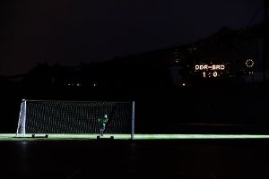 A football goal on a dark pitch. An illuminated sign shows West – East Germany 1:0.