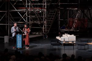 The curators Saba-Nur Cheema and Meron Mendel give their speech on stage at a lectern on which a banner with the festival logo is attached. In the background there is scaffolding from the set of the play “House”.