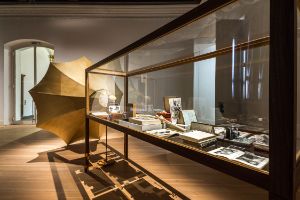 The photo shows the exhibition room, in which a glass case filled with multiple objects is placed, from which a big grammophone-horn extends.