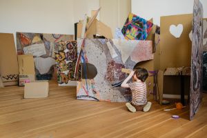 A child plays in a room with coloured cardboard and holds a cardboard tube to his eye like binoculars.