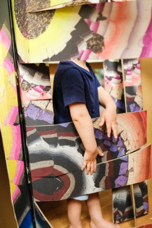 A child stands half-hidden behind a colourful cardboard wall and sticks out their hands.