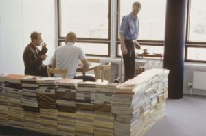 A table with chairs in front of a window. Around the table are boxes with paper catalogues on them. Two people are sitting on the table, one person is standing up.