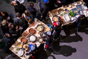 A shot from above shows two large tables with people gathered around them and various cooking utensils and ingredients for Pad Thai on the tables.