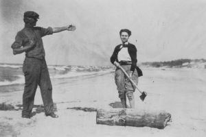 A man points to a woman staning with an axe over a tree trunk on the beach.