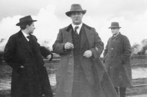 Three men in coats in front of a canal, the middle one looks into the camera