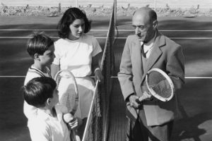 Mann im Anzug mit Tennisschläger spricht über ein Tennisnetz hinweg mit drei Kindern in Tennisbekleidung