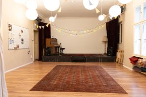 A room with a parquet floor covered with a carpet and a small stage with a piano on the back wall