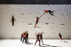 Performers are on and on top of a climbing wall, above which a performer lies on a rope stretched across the stage. 