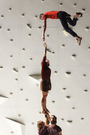 A performer reaches out from a rope to a female performer standing at the top of a human pyramid. 