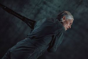 Lucinda Childs zieht an einem Seil. Die Choreografin trägt einen dunkelgrauen Overall und hat graue Haare.