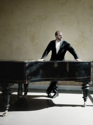 Kirill Gerstein stands behind a black grand piano.