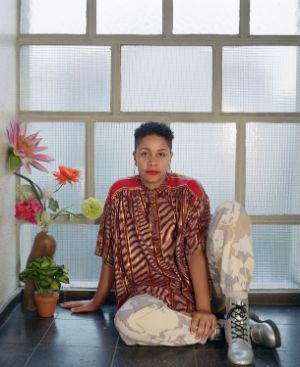 The picture shows a young person with short hair sitting in front of a window. She is wearing a patterned, loose-fitting top with red accents on the shoulders and camouflage-colored trousers. Her shoes are silver. On the left side of the picture is a vase with artificial flowers. The person is sitting on a dark floor in front of a large window with square panes. The window illuminates the scene with a soft, diffuse light.